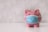 A pink piggy bank with white polka dots is wearing a blue surgical mask. The background appears to be a textured, light gray surface. The piggy bank is centered on a white surface, conveying a sense of concern or protection.