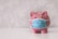A pink piggy bank with white polka dots is wearing a blue surgical mask. The background appears to be a textured, light gray surface. The piggy bank is centered on a white surface, conveying a sense of concern or protection.