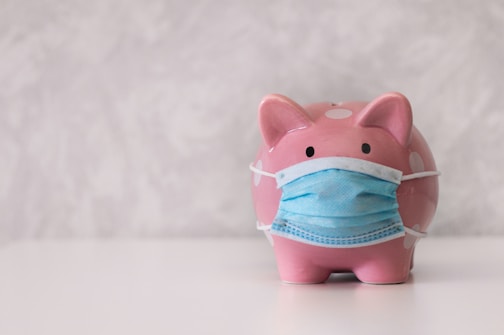 A pink piggy bank with white polka dots is wearing a blue surgical mask. The background appears to be a textured, light gray surface. The piggy bank is centered on a white surface, conveying a sense of concern or protection.