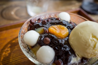 A dessert dish featuring a scoop of vanilla ice cream alongside sweet red beans, white mochi balls, and a bright yellow jelly-like center, served in a clear glass bowl.