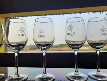 Several empty wine glasses are lined up on a reflective surface, with the logo of Sula Vineyards printed on each. The background shows a large window overlooking a serene landscape with greenery under a clear sky.