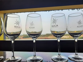 Several empty wine glasses are lined up on a reflective surface, with the logo of Sula Vineyards printed on each. The background shows a large window overlooking a serene landscape with greenery under a clear sky.