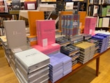 A table in a bookstore is stacked with various luxury brand coffee table books. The collection includes titles featuring high-end fashion brands such as Dior, Prada, Chanel, Louis Vuitton, and Yves Saint Laurent. The books are displayed in an organized manner, with some books upright and others stacked horizontally. They come in a variety of colors, including pink, gray, orange, blue, and black. Shelves filled with more books are visible in the background.