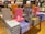 A table in a bookstore is stacked with various luxury brand coffee table books. The collection includes titles featuring high-end fashion brands such as Dior, Prada, Chanel, Louis Vuitton, and Yves Saint Laurent. The books are displayed in an organized manner, with some books upright and others stacked horizontally. They come in a variety of colors, including pink, gray, orange, blue, and black. Shelves filled with more books are visible in the background.