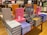 A table in a bookstore is stacked with various luxury brand coffee table books. The collection includes titles featuring high-end fashion brands such as Dior, Prada, Chanel, Louis Vuitton, and Yves Saint Laurent. The books are displayed in an organized manner, with some books upright and others stacked horizontally. They come in a variety of colors, including pink, gray, orange, blue, and black. Shelves filled with more books are visible in the background.