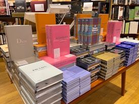 A table in a bookstore is stacked with various luxury brand coffee table books. The collection includes titles featuring high-end fashion brands such as Dior, Prada, Chanel, Louis Vuitton, and Yves Saint Laurent. The books are displayed in an organized manner, with some books upright and others stacked horizontally. They come in a variety of colors, including pink, gray, orange, blue, and black. Shelves filled with more books are visible in the background.