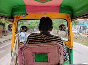 A view from inside a colorful auto-rickshaw, with striped fabric on the seat and vibrant colors around. The driver, seen from the back, wears a striped shirt. Two round side mirrors reflect the surroundings, capturing glimpses of a bicyclist and some greenery. The road ahead is visible, lined with protective barriers and greenery on one side.