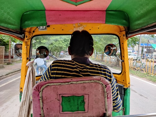 A view from inside a colorful auto-rickshaw, with striped fabric on the seat and vibrant colors around. The driver, seen from the back, wears a striped shirt. Two round side mirrors reflect the surroundings, capturing glimpses of a bicyclist and some greenery. The road ahead is visible, lined with protective barriers and greenery on one side.