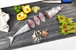 A cutting board with various cooking ingredients arranged neatly. There is a fish, cut into pieces, with its head and tail visible. Slices of lime, green and yellow bell peppers, garlic cloves, and a piece of ginger are also present. On one side, fresh lettuce leaves adorned with cherry tomatoes add a pop of color. A sharp knife with a blue handle rests at the top of the board.