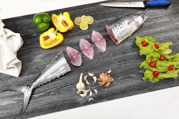 A cutting board with various cooking ingredients arranged neatly. There is a fish, cut into pieces, with its head and tail visible. Slices of lime, green and yellow bell peppers, garlic cloves, and a piece of ginger are also present. On one side, fresh lettuce leaves adorned with cherry tomatoes add a pop of color. A sharp knife with a blue handle rests at the top of the board.