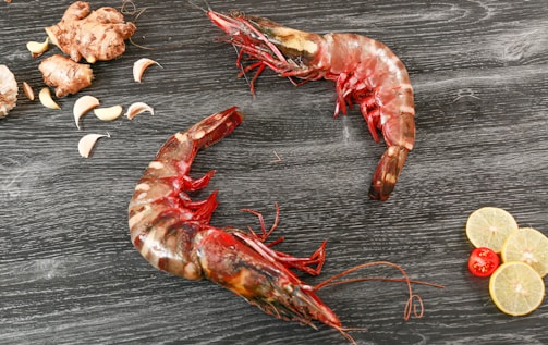 Two raw tiger prawns are placed on a dark wooden surface with scattered garlic cloves, ginger, lemon slices, and a red cherry tomato.