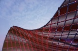 A dynamic architectural structure with a curved, mesh-like surface is partially illuminated by small red lights. The sky in the background is a pale blue, creating a stark contrast with the metal framework.