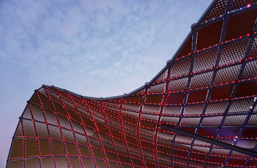 A dynamic architectural structure with a curved, mesh-like surface is partially illuminated by small red lights. The sky in the background is a pale blue, creating a stark contrast with the metal framework.