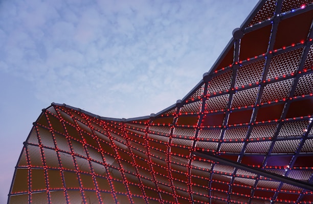 A dynamic architectural structure with a curved, mesh-like surface is partially illuminated by small red lights. The sky in the background is a pale blue, creating a stark contrast with the metal framework.