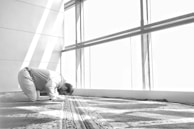 A person kneels in prayer on a patterned carpet in a bright room. Sunlight streams through a large window, casting clear shadows.