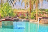 A tranquil swimming pool is surrounded by lush palm trees and vibrant flowering bushes. Stone and tile elements border the pool, creating a luxurious atmosphere. A sculpture of a human figure stands amongst the plants, adding an artistic touch. The background features a view of distant mountains under a clear blue sky, enhancing the serene setting.