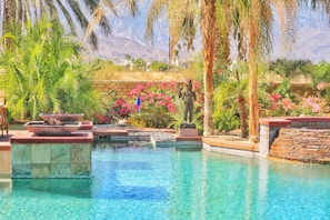 A tranquil swimming pool is surrounded by lush palm trees and vibrant flowering bushes. Stone and tile elements border the pool, creating a luxurious atmosphere. A sculpture of a human figure stands amongst the plants, adding an artistic touch. The background features a view of distant mountains under a clear blue sky, enhancing the serene setting.