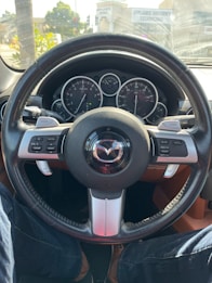 A car dashboard with a steering wheel bearing the Mazda logo. Instrument gauges showing speed and RPMs are visible, and the steering wheel has buttons for various controls. The image includes a view through the windshield of an urban street with some foliage and buildings.