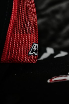 A close-up of a red mesh fabric with a sewn patch featuring a letter. The background is dark, adding contrast to the vivid red color.
