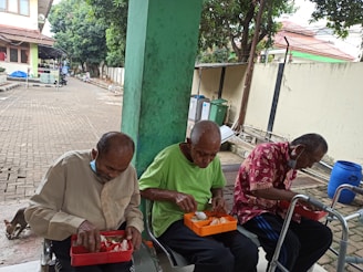 Three elderly men are sitting on benches outside, each with a bento-style container of food on their laps. They are focused on eating. Behind them is a paved walkway, various trees, and residential buildings. One man wears a beige shirt, another a green shirt, and the third a red patterned shirt. They each have a face mask pulled down under their chin.