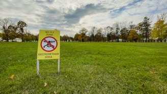 A grassy open field with a yellow sign prohibiting drone flights. The sign features a pictogram of a drone with a red circle and a line through it, accompanied by text in both English and another language. The background comprises trees with autumn foliage under a cloud-filled sky.