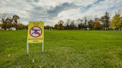 A grassy open field with a yellow sign prohibiting drone flights. The sign features a pictogram of a drone with a red circle and a line through it, accompanied by text in both English and another language. The background comprises trees with autumn foliage under a cloud-filled sky.