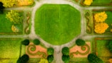 An aerial view of a landscaped garden featuring a symmetrical layout with a large central grassy area, surrounded by pathways and decorative patterns of trimmed hedges. Trees with autumnal foliage provide contrast against the vibrant green grass.