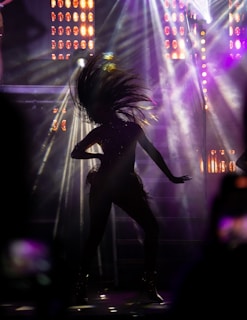 A silhouette of a person captured mid-dance with their hair dramatically tossed back. The background is illuminated with vibrant stage lights and beams, creating an energetic and dynamic atmosphere.