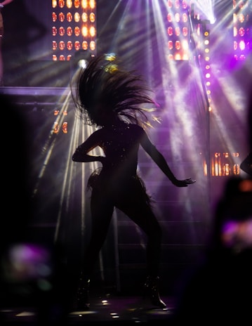 A silhouette of a person captured mid-dance with their hair dramatically tossed back. The background is illuminated with vibrant stage lights and beams, creating an energetic and dynamic atmosphere.
