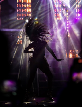 A silhouette of a person captured mid-dance with their hair dramatically tossed back. The background is illuminated with vibrant stage lights and beams, creating an energetic and dynamic atmosphere.