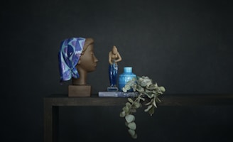 A still life setup features a realistic sculpture of a woman's head adorned with a colorful, patterned headscarf in shades of blue and purple. Next to it, a small ceramic sculpture of a human figure stands, alongside a blue vase and a greenish branch of eucalyptus resting on a book. The scene is set against a dark, muted backdrop, creating a calm and serene composition.