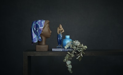 A still life setup features a realistic sculpture of a woman's head adorned with a colorful, patterned headscarf in shades of blue and purple. Next to it, a small ceramic sculpture of a human figure stands, alongside a blue vase and a greenish branch of eucalyptus resting on a book. The scene is set against a dark, muted backdrop, creating a calm and serene composition.