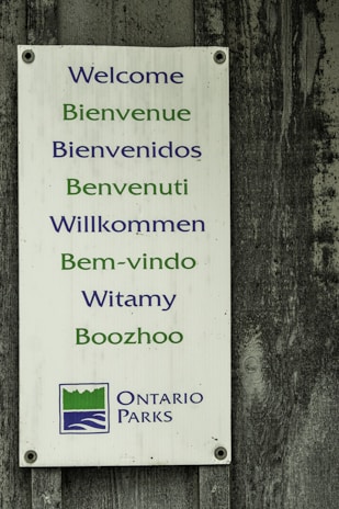 A rectangular sign with welcoming messages in multiple languages including English, French, Spanish, Italian, German, Portuguese, Polish, and Anishinaabemowin. The sign has blue and green text and features the Ontario Parks logo at the bottom. It is mounted on a weathered wooden surface.