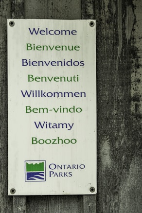 A rectangular sign with welcoming messages in multiple languages including English, French, Spanish, Italian, German, Portuguese, Polish, and Anishinaabemowin. The sign has blue and green text and features the Ontario Parks logo at the bottom. It is mounted on a weathered wooden surface.