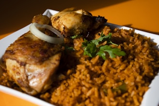A close-up of a dish featuring golden-brown grilled chicken pieces served with a side of vibrant orange rice garnished with fresh green herbs. A ring of raw onion is placed on top of the chicken.