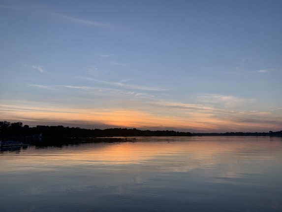 A serene lake view at sunset, with soft reflections of the glowing orange and pink hues on the calm water surface. The sky transitions from a deep blue at the top to a warm orange near the horizon, with silhouetted trees lining the distant shore.