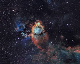 A colorful and detailed image of a nebula set against a vast backdrop of space. The center of the nebula is brightly highlighted with shades of orange, red, and blue. Dark dust lanes create intricate patterns within the glowing gas, while numerous stars dot the surrounding space, adding to the depth and complexity of the scene.