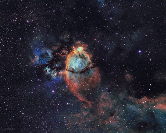 A colorful and detailed image of a nebula set against a vast backdrop of space. The center of the nebula is brightly highlighted with shades of orange, red, and blue. Dark dust lanes create intricate patterns within the glowing gas, while numerous stars dot the surrounding space, adding to the depth and complexity of the scene.