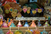 A display of colorful handcrafted ceramic ornaments, including hearts with inspirational words, circular designs with floral patterns, and whimsical cone-shaped figures. The arrangement is vibrant and artistic, with various shapes and messages promoting positivity.