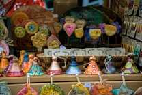 A display of colorful handcrafted ceramic ornaments, including hearts with inspirational words, circular designs with floral patterns, and whimsical cone-shaped figures. The arrangement is vibrant and artistic, with various shapes and messages promoting positivity.