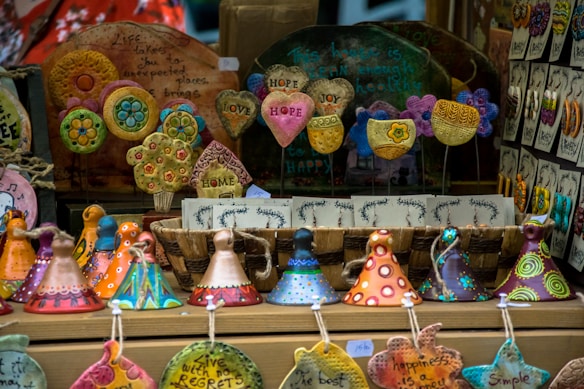 A display of colorful handcrafted ceramic ornaments, including hearts with inspirational words, circular designs with floral patterns, and whimsical cone-shaped figures. The arrangement is vibrant and artistic, with various shapes and messages promoting positivity.