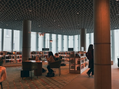 A modern library interior with large windows allowing natural light to illuminate the space. The ceiling has an intricate geometric pattern, and there are several tall pillars throughout the room. People are seen working on laptops or reading at tables, surrounded by shelves filled with books.