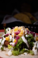 A close-up view of a taco filled with ingredients like lettuce, corn, shredded cheese, pickled onions, and a drizzle of white sauce on top. The taco shell is partially visible, with additional tacos blurred in the background.