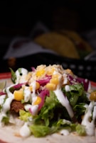 A close-up view of a taco filled with ingredients like lettuce, corn, shredded cheese, pickled onions, and a drizzle of white sauce on top. The taco shell is partially visible, with additional tacos blurred in the background.