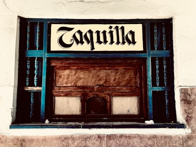 A vintage ticket booth with wooden panels, painted with teal accents. The window is encased in a grid of teal rails, and a sign above reads 'Taquilla' in a bold, blackletter font. The surrounding wall is textured and painted white.