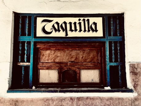 A vintage ticket booth with wooden panels, painted with teal accents. The window is encased in a grid of teal rails, and a sign above reads 'Taquilla' in a bold, blackletter font. The surrounding wall is textured and painted white.