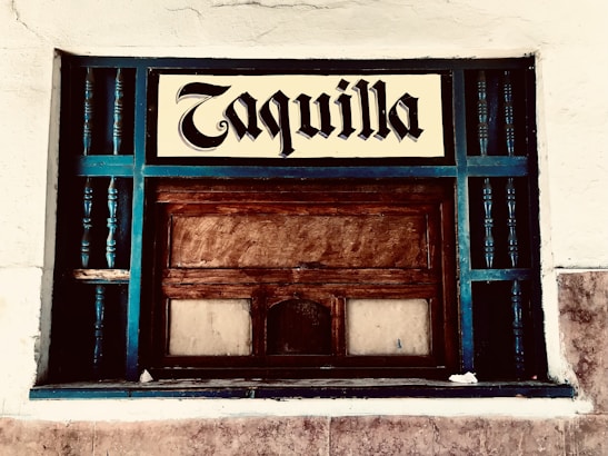 A vintage ticket booth with wooden panels, painted with teal accents. The window is encased in a grid of teal rails, and a sign above reads 'Taquilla' in a bold, blackletter font. The surrounding wall is textured and painted white.