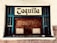 A vintage ticket booth with wooden panels, painted with teal accents. The window is encased in a grid of teal rails, and a sign above reads 'Taquilla' in a bold, blackletter font. The surrounding wall is textured and painted white.