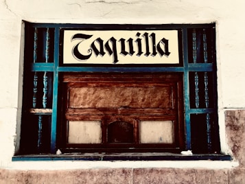 A vintage ticket booth with wooden panels, painted with teal accents. The window is encased in a grid of teal rails, and a sign above reads 'Taquilla' in a bold, blackletter font. The surrounding wall is textured and painted white.