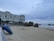 A seaside landscape with a large, elegant white building along a beach promenade. In the foreground, a blue car is precariously positioned halfway off a low wall leading down to the sandy beach. To the right, rock formations are present along the shore, and the ocean stretches into the distance under a cloudy sky. Several people are walking along the promenade and the beach.